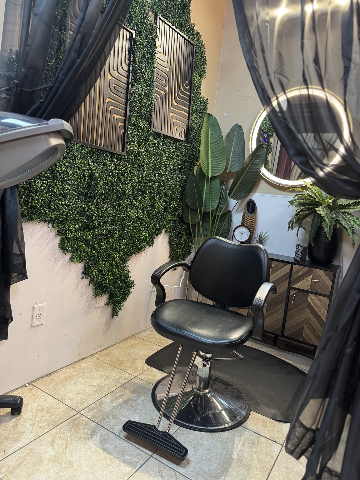 Angled view of a styling chair, round mirror, decorative greenery wall, and privacy curtains